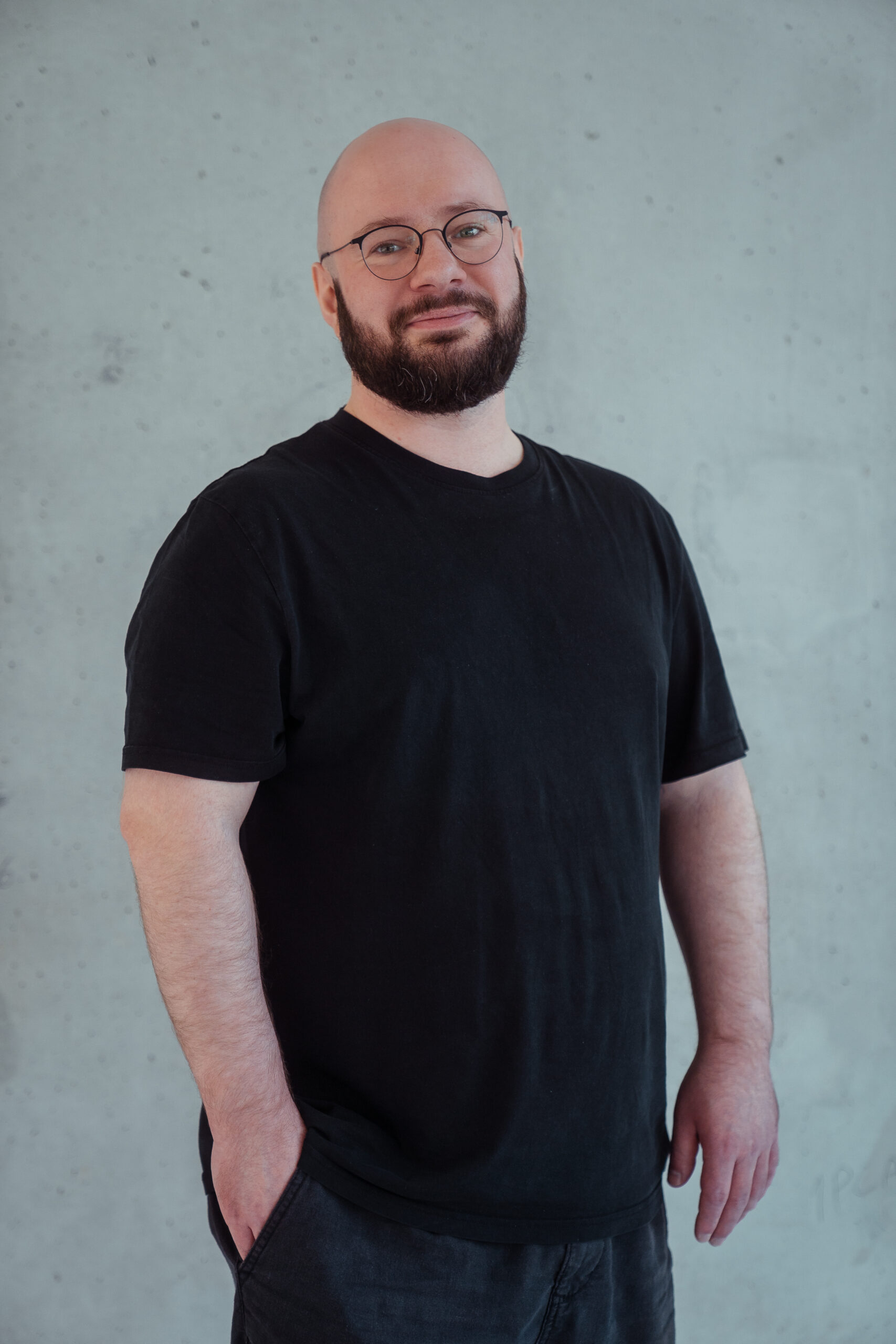 Porträt des Mitarbeiters Henry, der im Bereich digitale Prozesse und Marketing bei DREILAUT tätig ist. Er steht vor einem hellgrauen Hintergrund, hat eine Glatze, trägt eine dünne, runde Brille, ein schwarzes T-Shirt mit einer dunklen Hose und schaut freundlich in die Kamera.