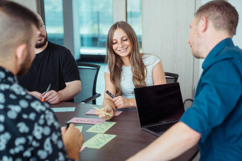 Momentaufnahme eines Workshops. Die Teilnehmenden sitzen in einem Meetingraum an einem großen Tisch, auf dem sich neben eines Laptops viele beschriebene, bunte Karteikarten befinden.
