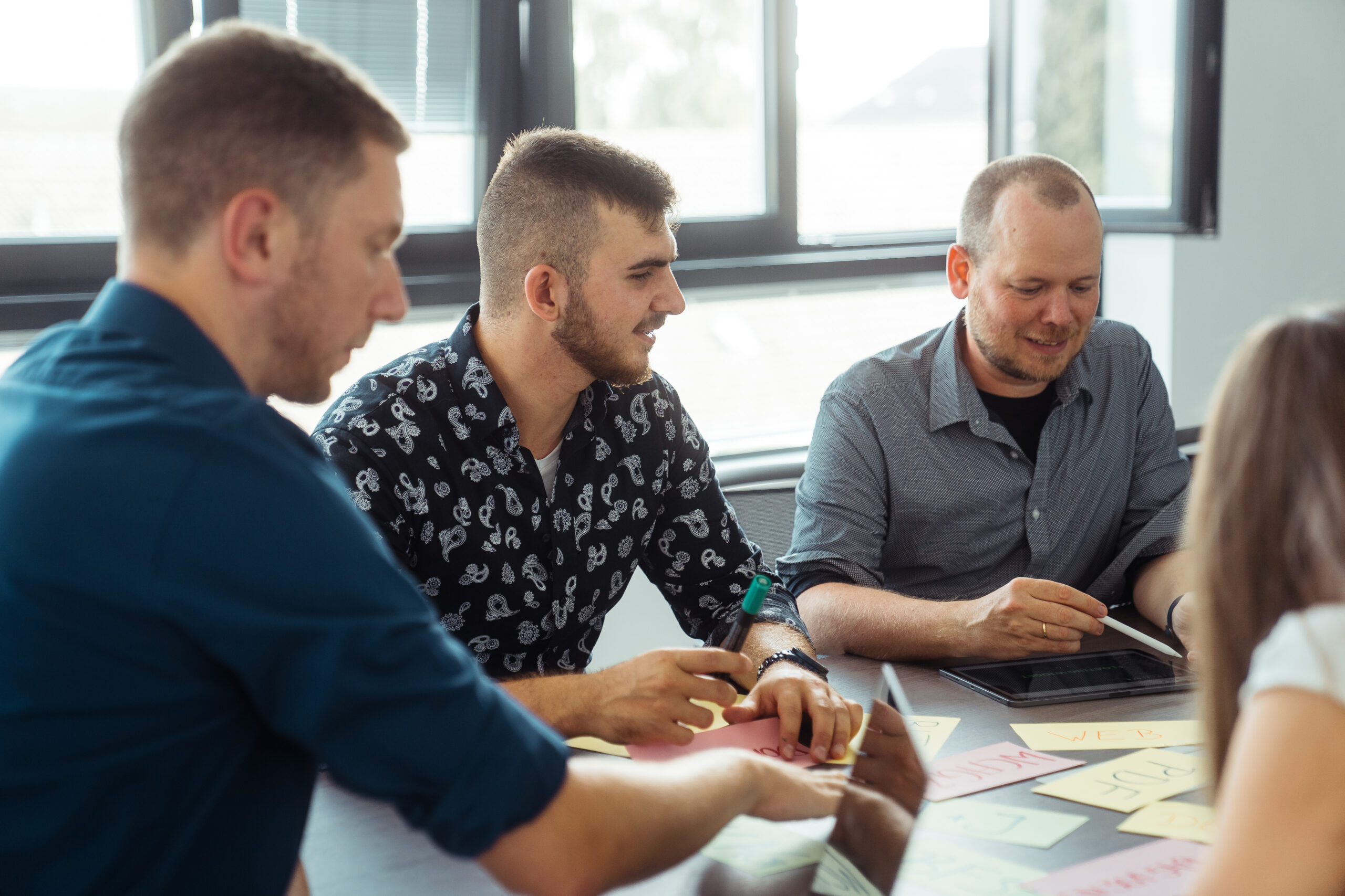 Momentaufnahme eines Workshops, bei dem die Mitarbeiter Sinan Fischer, Daniel Petermann-Fallis, Emily von Känel und der Geschäftsführer Christoph Konitzer in einem Meetingraum an einem großen Tisch sitzen, auf dem sich neben eines Laptops und eines iPads viele beschriebene, bunte Karteikarten befinden. Der Geschäftsführer Christoph Konitzer schaut auf die Karteikarten, währenddessen der Mitarbeiter Sinan Fischer lächelnd zu Daniel Petermann-Fallis schaut, der mit einem Stift etwas auf seinem iPad zeigt, das vor ihm liegt.