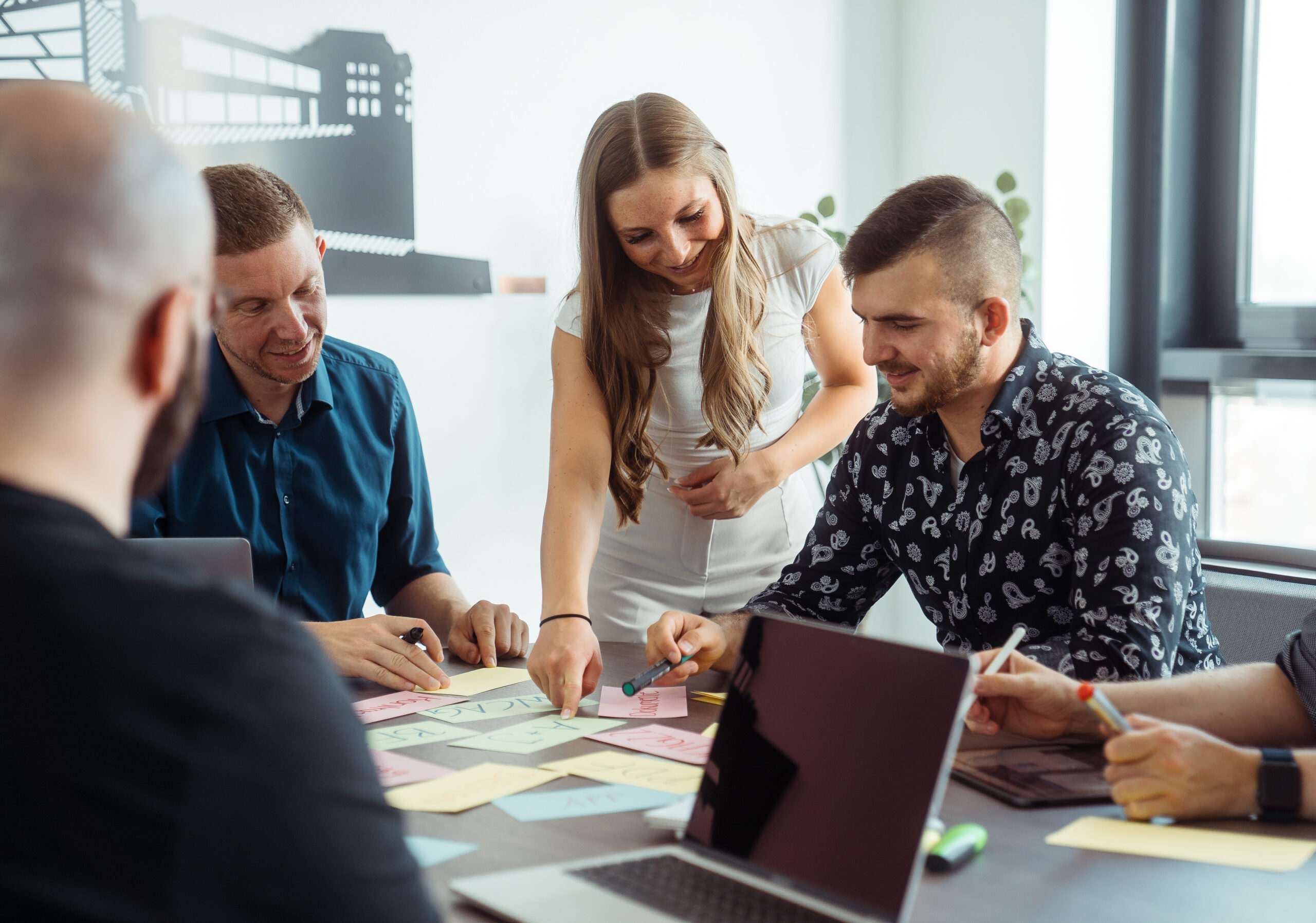 Momentaufnahme eines Workshops, bei dem die fröhlich lächelnden Teilnehmenden in einem Meetingraum an einem großen Tisch sitzen, auf dem sich neben beschriebenen, bunten Karteikarten, ebenfalls Stifte sowie ein Laptop befinden.