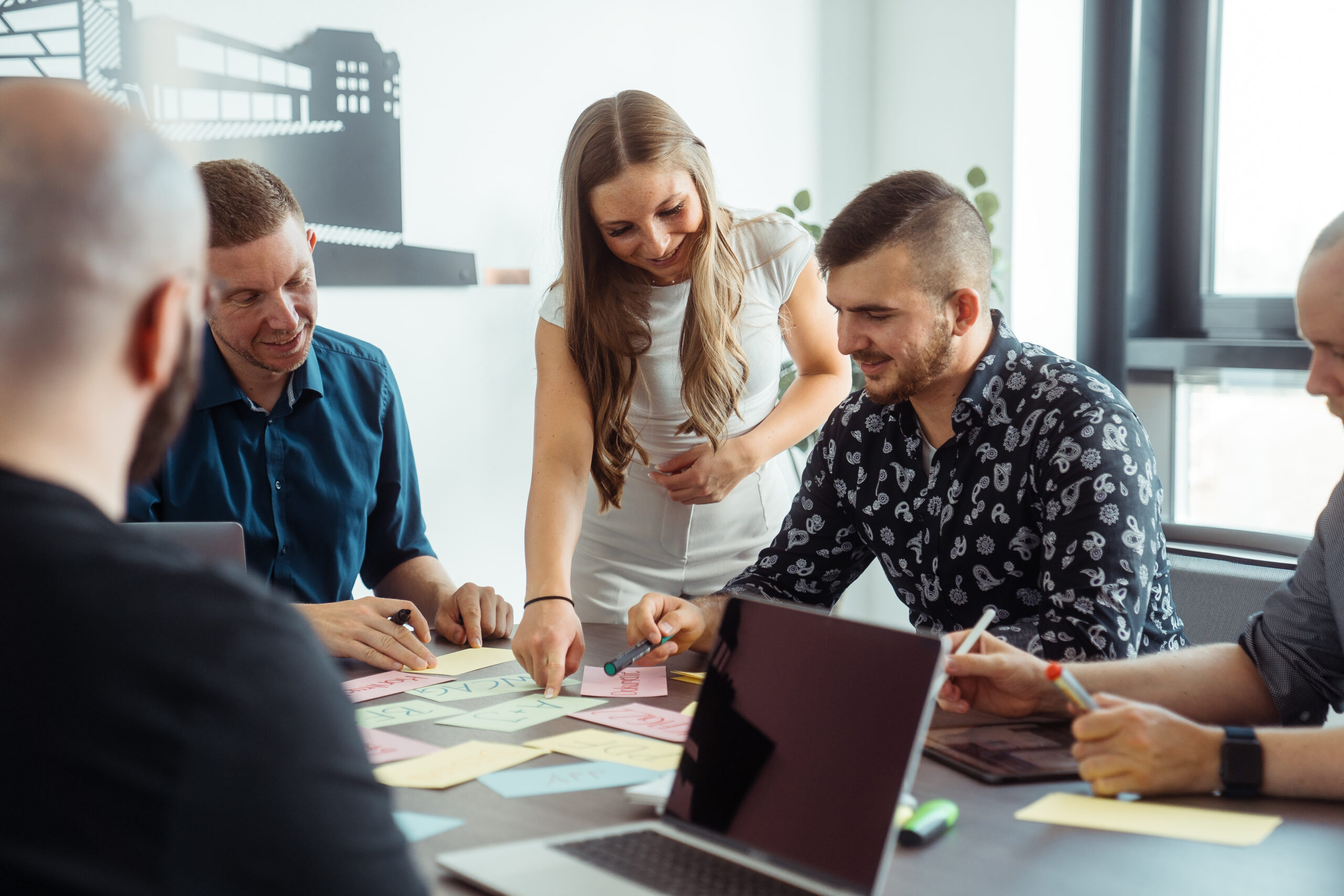 Momentaufnahme eines Workshops, bei dem die fröhlich lächelnden Mitarbeiter Daniel Petermann-Fallis, Henry Ganschof, Sinan Fischer und der Geschäftsführer Christoph Konitzer in einem Meetingraum an einem großen Tisch sitzen, auf dem sich neben beschriebenen, bunten Karteikarten, ebenfalls Stifte sowie ein iPad und ein Laptop befinden. Die Mitarbeiterin Emily von Känel steht zwischen Christoph Konitzer und Sinan Fischer und lehnt sich leicht über den Tisch, um auf eine grüne Karteikarte mit der Aufschrift "A+J" zu verweisen, auf die der Blick aller Mitarbeiter gerichtet ist.