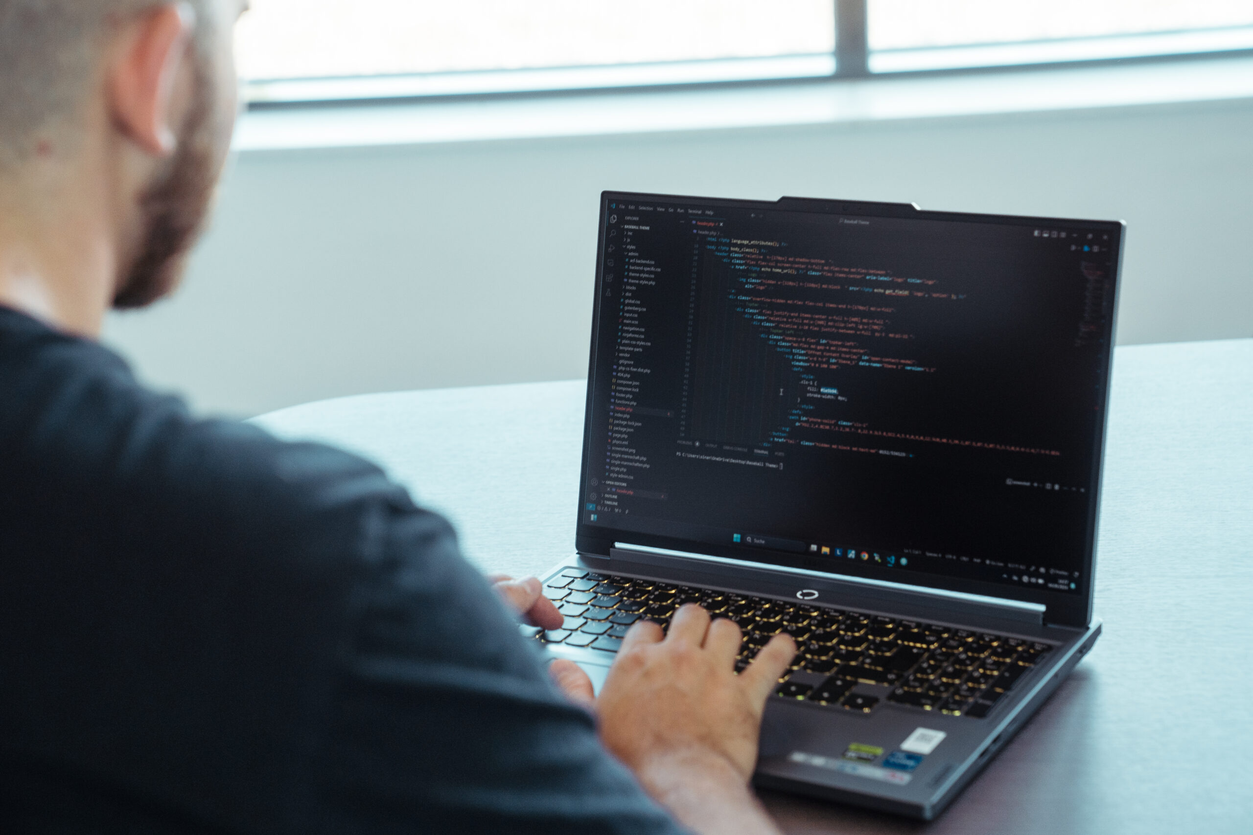 Ein Mann von hinten, der an einem Tisch sitzt und an einem Laptop tippt. Auf dem Bildschirm des Laptops stehen HTML-Codes, die in einem Programmier-Programm geöffnet sind.
