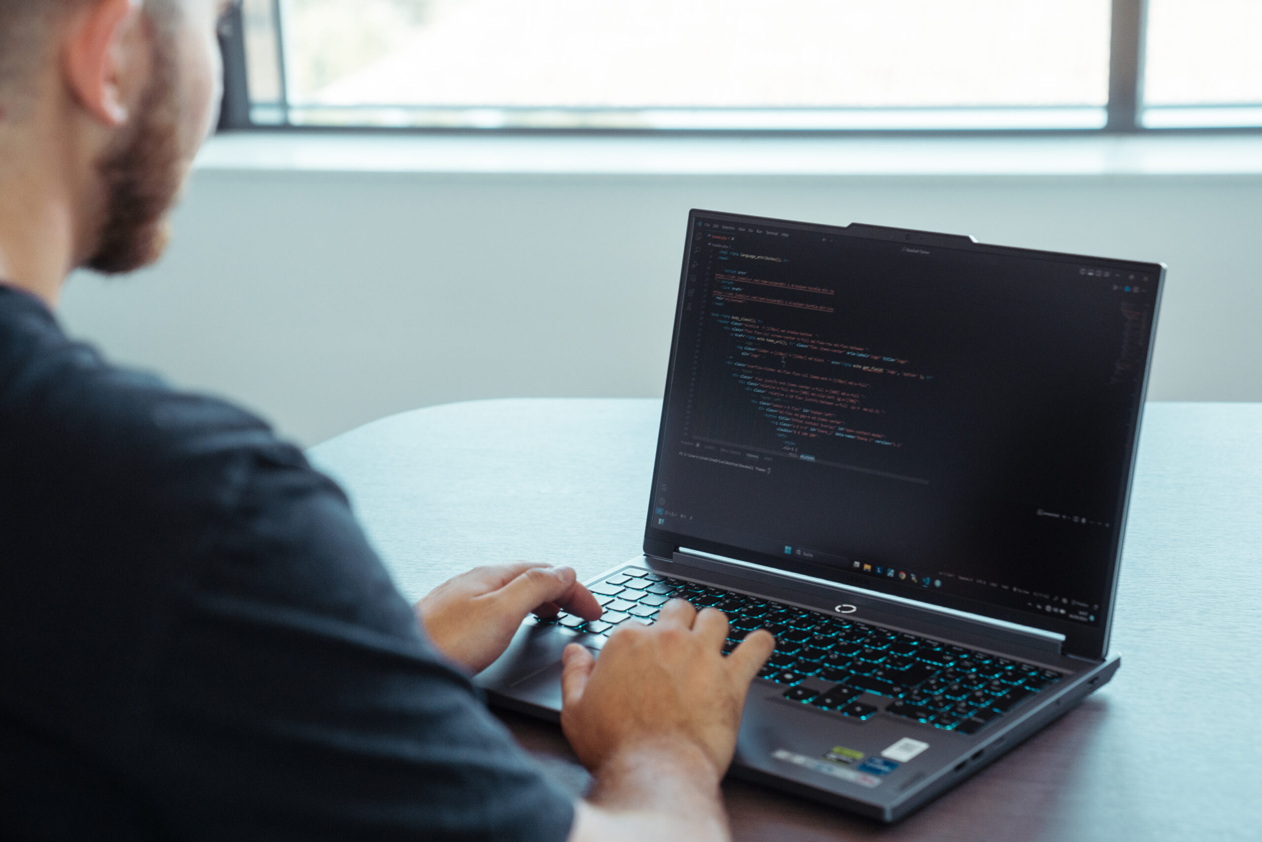 Ein Mann von hinten, der an einem Tisch sitzt und an einem Laptop tippt. Auf dem Bildschirm des Laptops stehen HTML-Codes, die in einem Programmier-Programm geöffnet sind.